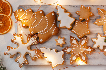 Delicious homemade gingerbread cookies with white icing. Christmas lights, branch of fir tree, dry orange slices. White wooden background, flat lay, top view. Festive holiday atmosphere, family time