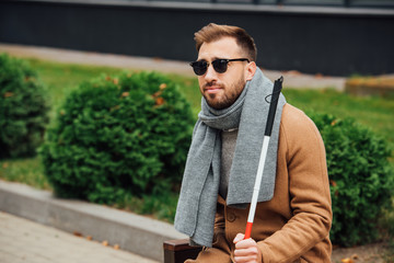 Blind man in sunglasses holding walking stick on bench