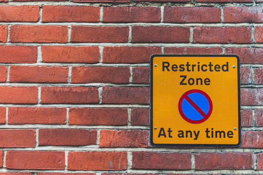 picture of a restricted zone sign on a old orange brick wall. Yellow sign, black letters, blue circle crossed out.