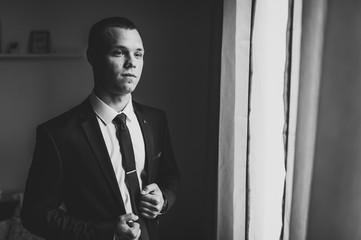 Portrait of a young attractive handsome businessman stylish serious man in a suit standing on the background room near window. Black and white photo.