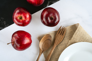 Fresh juicy red apples lie on black and white marble surface with wooden spoon and fork, empty white plate on a sackcloth. Top view flat lay composition.