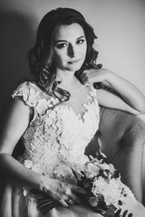 Portrait of a beautiful young bride with wedding bouquet of flower on retro armchair. Tender emotion on the face. Black and white photo.