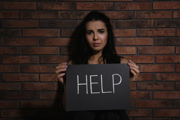 Abused young woman with sign HELP near brick wall. Domestic violence concept