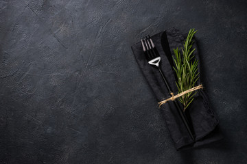 Black cutlery and napkin with a sprig of rosemary on black stone table top view.