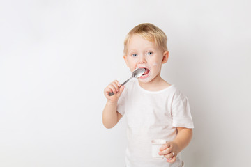 portrait of a blond blue-eyed little caucasian cute boy eating a yogurt with a spoon on a white...