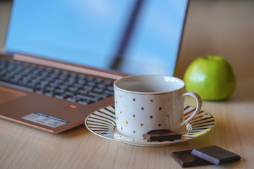 Cup of coffee on the wooden table with notebook on the background. Piece of black chocolate is on the saucer, break time in the office for refreshment