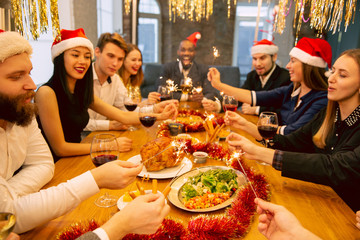 Happy co-workers celebrating while company New Year and Christmas party. Young caucasian people in celebrate attire with sparklers talk, drink wine, cheers. Concept of office culture, teamwork, winter