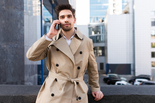 Young stylish man in trench coat talking on cellphone confidently looking in camera on street