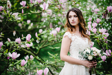 Portrait of bride with a wedding bouquet standing on background nature of purple flowers of magnolia and greens. outdoors. looking sideways. upper half length.