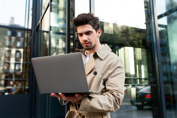 Young stylish man in trench coat confidently working on laptop on street