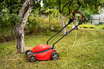 Red lawn mower outdoor in the backyard. green grass and fruit trees background. Gardening concept.