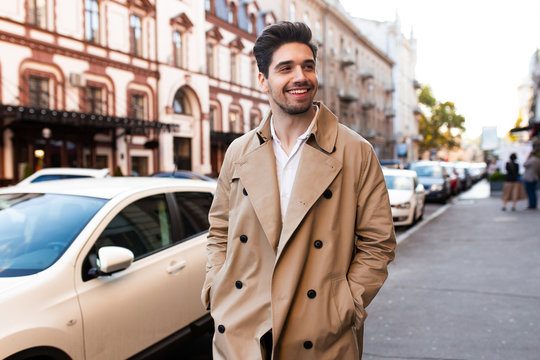 Young attractive smiling stylish man in trench coat happily walking through street