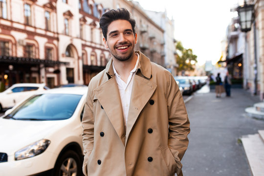 Young handsome smiling stylish man in trench coat joyfully walking through street
