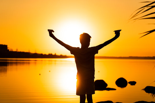 Little Kid Holds Two Paper Planes In Spread Hands And Plays At Sunset On The Lake Background. Time Outdoor. Black Silhuette Against Sundown