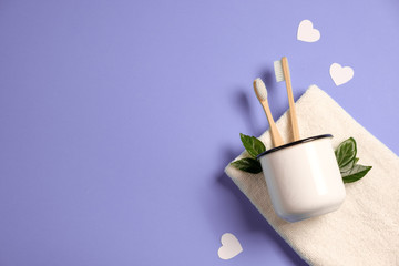 Two wooden bamboo eco friendly toothbrushes in metal cup, green leaf, white heart and towel on purple background. Eco friendly, reuse and valentines day concept. Flat lay, top view, copy space - Powered by Adobe