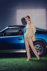 attractive and stylish woman in suit with hands in pocket standing near retro car