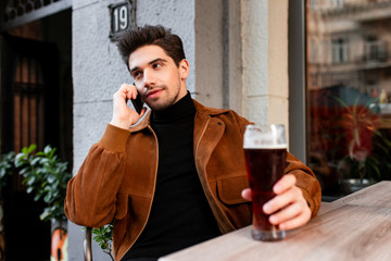 Young casual man talking on cellphone sitting with beer in cafe on street