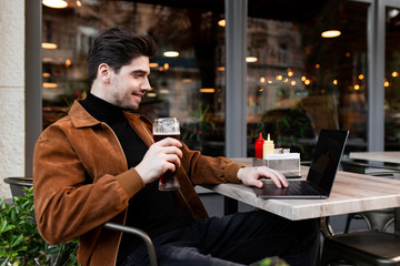 Young handsome casual businessman happily working on laptop while drinking beer in cafe on street