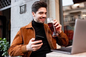 Young attractive casual man with cellphone happily drinking beer working on laptop in cafe on street