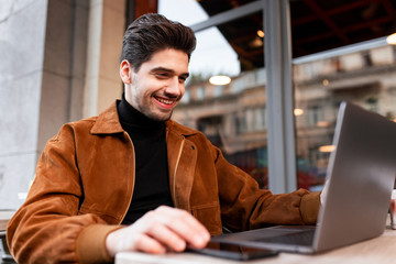 Young handsome casual businessman joyfully working on laptop in cafe on street