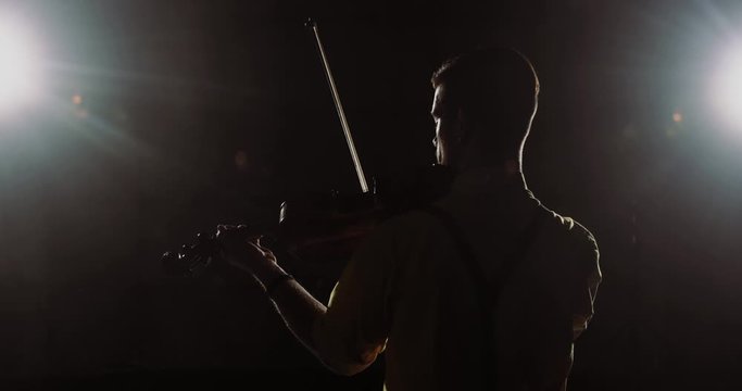 Popular musician playing solo on his his instrument violin on smoked black background. Man professional violinist performs from stage in light of spotlight, back view. Classical music player.