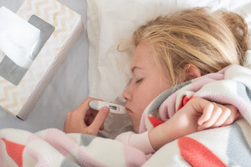 Sick teenage girl lying on the bed covered with blanket with thermometer in her mouth ro measure body temoerature and box of rissue on side