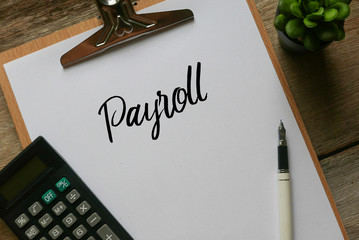 Top view of plant,calculator,pen and clipboard with white paper written with Payroll on wooden...