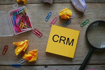 Top view of colorful paper clips magnifying glass,trash paper and yellow sticky note written with CRM (Customer Relationship Management) on wooden background.