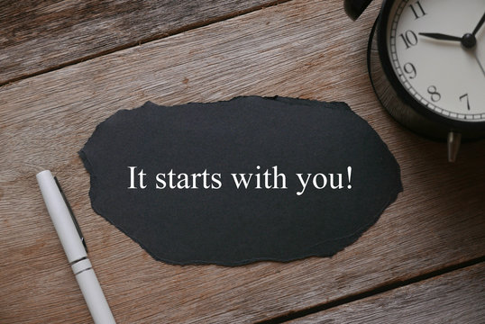 Top view and selective focus of clock,pen and a piece of black paper written with It Starts With You! on wooden background.