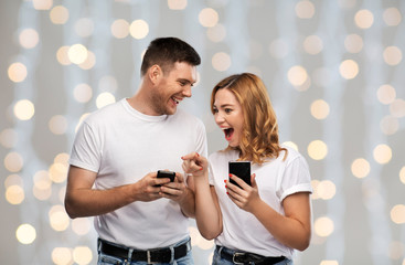 technology and people concept - happy couple in white t-shirts with smartphones over festive lights...