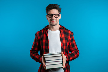European student in red plaid shirt on blue background in studio holds stack of university books from library. Guy smiles, he is happy to graduate.