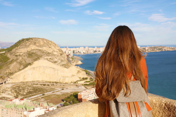 Fototapeta na wymiar Back view of traveler girl enjoying view of Alicante city from Santa Barbara castle, Spain. Young female backpacker relaxing in her travel in southern Europe.