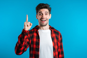 Portrait of young thinking pondering man in red having idea moment pointing finger up on blue...