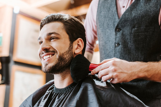 barber holding cosmetic brush near face of happy bearded man