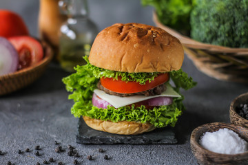 Photo of fresh burger on wooden cutting board on dark background..Homemade hamburger with beef, onion, tomato, lettuce and cheese. Homemade fast food. Dark textured background. Copy space. Image.