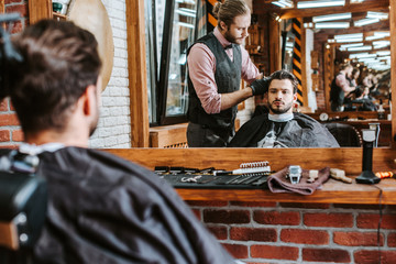 selective focus of handsome barber styling hair of man near mirror in barbershop