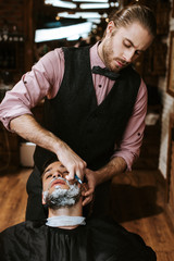 stylish barber shaving bearded man with shaving cream on face