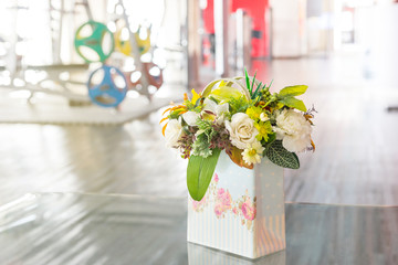 Decoration artificial flower in colorful vintage style vase on table with blurred of fitness gym background. Beautiful white roses flower bouquet in a box in fitness center, background or copy space.