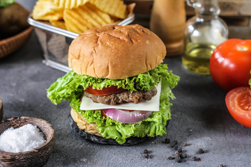Photo of fresh burger on wooden cutting board on dark background..Homemade hamburger with beef, onion, tomato, lettuce and cheese. Homemade fast food. Dark textured background. Copy space. Image.