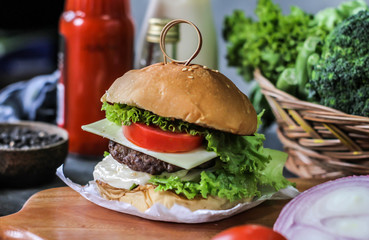 Photo of fresh burger on wooden cutting board on dark background..Homemade hamburger with beef, onion, tomato, lettuce and cheese. Homemade fast food. Dark textured background. Copy space. Image.