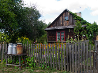 Stary drewniany dom z niewielkim ogrodem. Drewniany płot z sztachet i kanki, zbiorniki na mleko