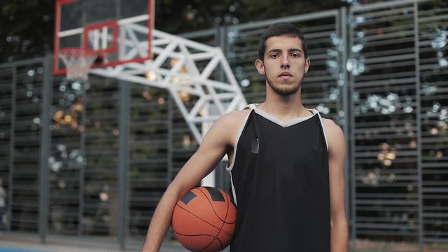 Portrait of Young Slim Fit Serious Caucasian Guy in Black Singlet Holding Ball Standing at Street Basketbal Court and Looking to Camera. Healthy Lifestyle and Sport Concept.