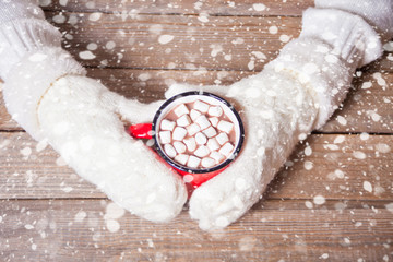 Woman's hand in white mittens holding red mug of Christmas cocoa with marshmallows.