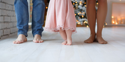 Parts of the body. Feet of dad, mom and baby. The baby takes the first steps with the help of parents, close up. Parents and their little daughter stand bare feet on a white floor against the backgrou