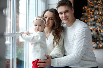 A happy family, father, mother and little daughter, spend time together on Christmas evening in a bright room indoors against a background of a beautifully decorated Christmas tree with garlands and g