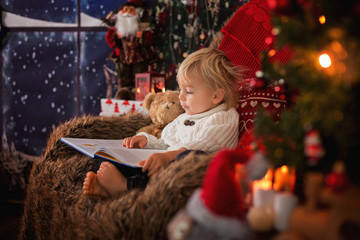 Cute toddler boy, sitting around christmas tree in cozy chair, reading book