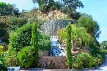 Naklejka premium A small decorative waterfall on a cliff. Palm trees, cypress trees and southern evergreens. Landscaping.
