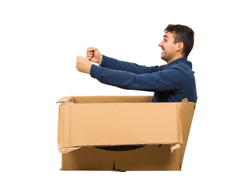Full length side view of childish man sitting inside a cardboard box pretending to drive a new car isolated over white background. Joyful guy dreaming of buying a personal vehicle.