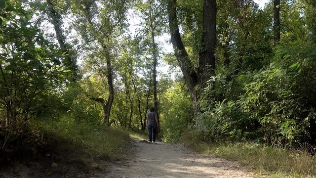 4K view of a woman walking along a path among green trees and bushes. Woman moves deep into the forest