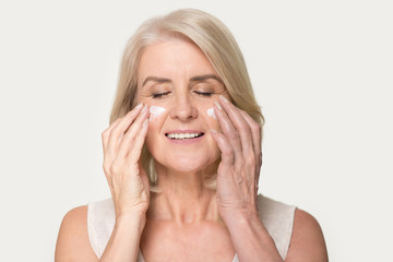 Smiling mature woman applying rejuvenating cream on her face skin